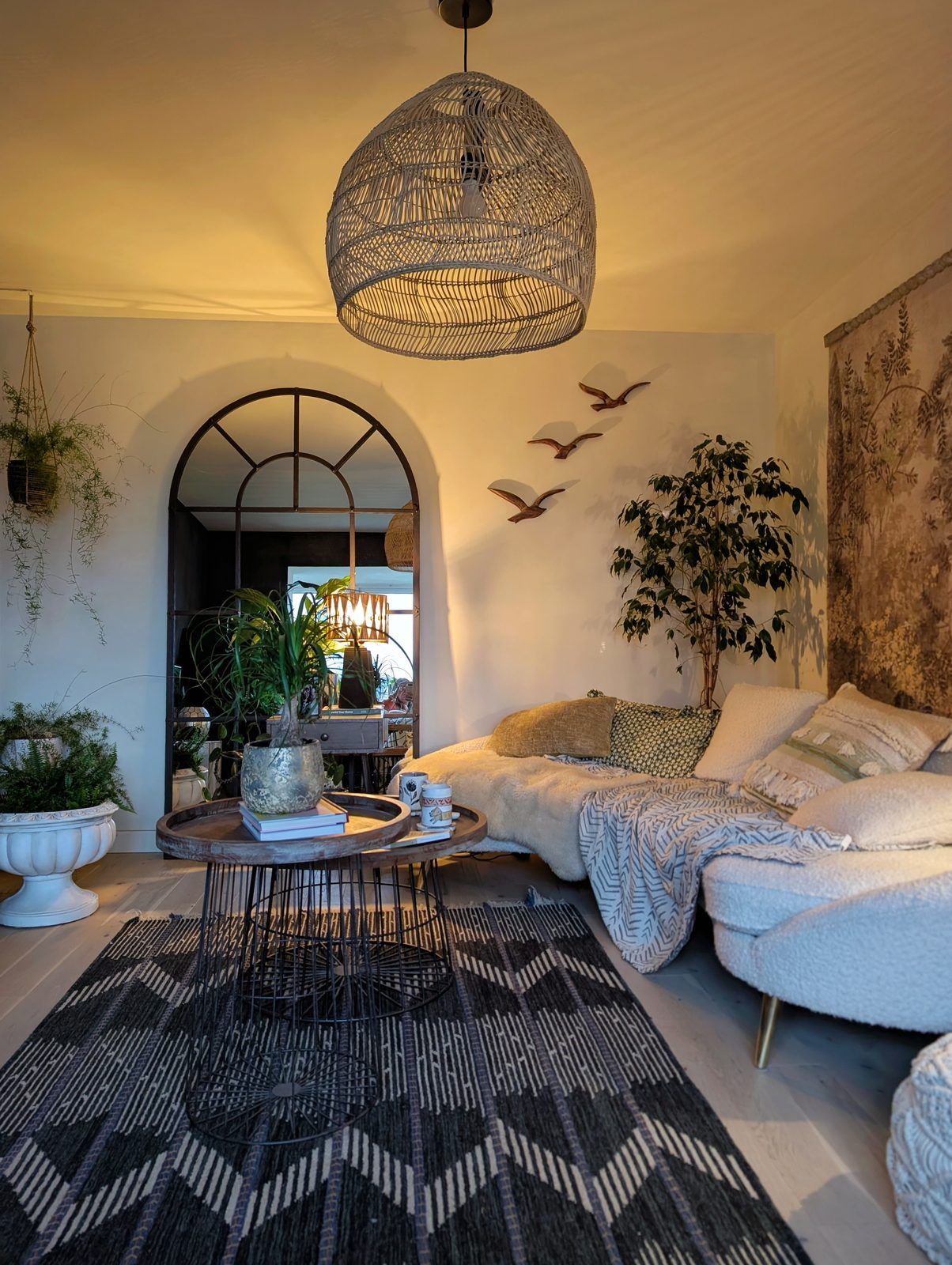 Golden-hour living room — rattan pendant, curved bouclé sofa, layered cushions, arched mirror, plants and patterned rug
