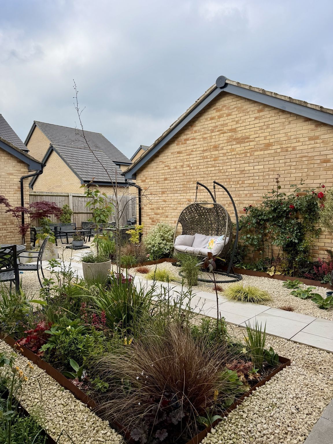 Contemporary garden built from a new-build plot — hanging chair, layered planting beds, gravel and paving, with climbing roses on the brick walls.